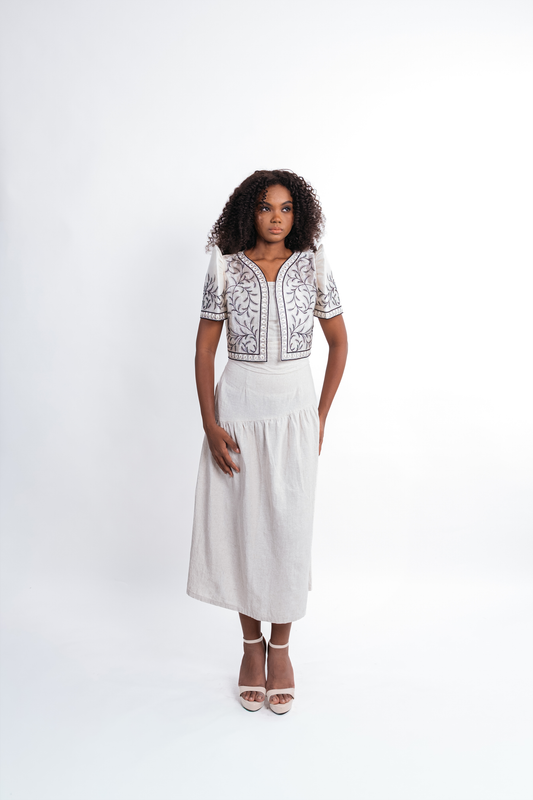 A woman modeling a white midi dress with a bolero top featuring black and gray floral embroidery, paired with white high-heeled sandals, against a plain white studio background.